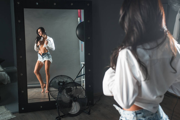 young girl with dark hair in denim shorts and in a white shirt in a dark room standing by the mirror looking at herself - Foto, imagen