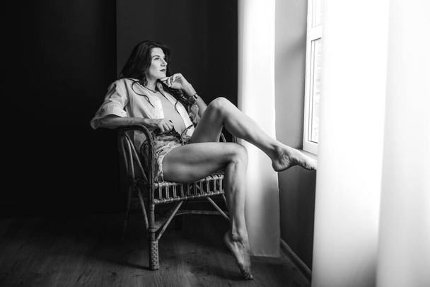 a young girl with dark hair in denim shorts and a white shirt in a dark room sitting on a chair by the window conceived,  black and white photo - Photo, image