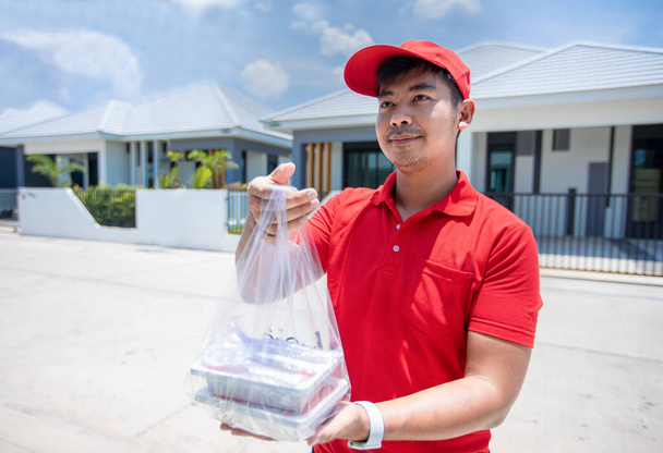 Asiatische Lieferdienste tragen eine rote Uniform mit roter Mütze und hantieren mit Lebensmittelboxen in Plastiktüten, die sie vor dem Haus an den Kunden verteilen. Online-Einkauf und Express-Lieferung   - Foto, Bild