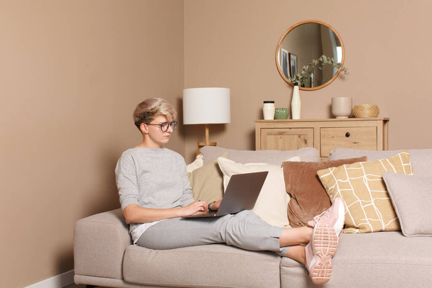 Young serious female freelancer working or learning from home. Full body of pretty intelligent woman 30s in casual clothes and glasses sitting on a sofa indoors typing on a laptop - Valokuva, kuva