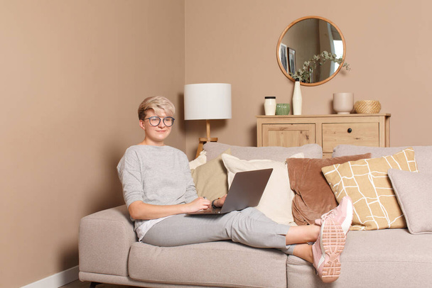 Młoda szczęśliwa kobieta freelancerka pracująca lub ucząca się w domu podczas samoizolacji. Portret bardzo inteligentnej kobiety po trzydziestce w luźnych ubraniach i okularach, siedzącej na kanapie w domu i piszącej na laptopie, uśmiechającej się.. - Zdjęcie, obraz