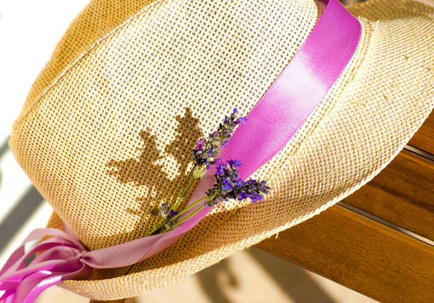 Ανοιχτό ψάθινο καπέλο σε ξύλινο καφέ τραπέζι. Λιλακ-ροζ κορδέλα στο καπέλο της γυναίκας. μπουκέτο από ξερή λεβάντα σε ρουστίκ ψάθινο καλάθι. - Φωτογραφία, εικόνα