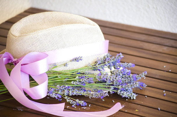 Lichte strohoed op houten bruine tafel. Lilac-roze lint op hoed van dameshoed. boeket droge lavendel in rustieke rieten rieten rieten mand. - Foto, afbeelding