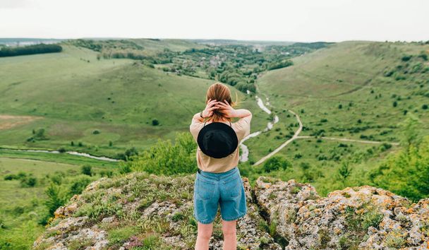 Mujer Hipster está de pie con su espalda a una roca y mira a un hermoso paisaje verde con un río y un valle. El turista se encuentra en una colina y admira el paisaje. Turismo doméstico
 - Foto, imagen