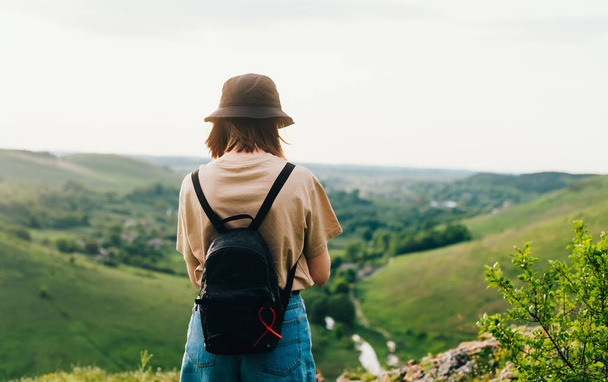 Rücken eines Touristenmädchens steht auf einem Berg mit einer schönen grünen Landschaft. Hipster-Frau in Freizeitkleidung und mit Rucksack steht am Rande des Berges. - Foto, Bild
