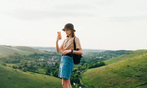 Attractive hipster girl in stylish clothes stands on top of a mountain in Tovtry and uses a smartphone. Girl tourist stands on a rock in beautiful Ukrainian landscape and takes photos on a smartphone - Photo, Image