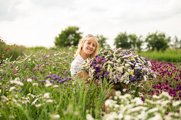 Usmívající se dospělá žena sedící v květinové zahradě s velkou kyticí květin v rukou a dívající se na kameru se šťastnou tváří. Šťastná žena drží řezané květiny pěstované v zahradním lůžku v chatě - Fotografie, Obrázek