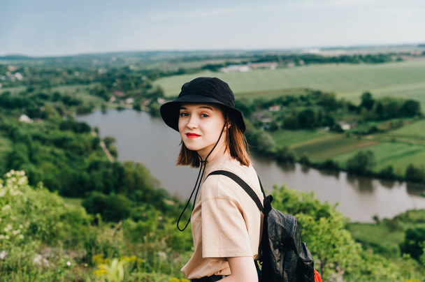 Positivo turista ragazza in abiti casual in piedi su una collina sullo sfondo del lago e guardando la fotocamera con un sorriso sul viso. Sorridente hipster ragazza è impegnata nel turismo domestico - Foto, immagini
