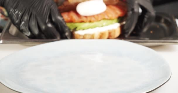 Close-up of cook's hands in gloves serving a delicious croissant with cheese, avocado, slices of salmon and poached egg on a plate. Tasty and healthy breakfast. - Záběry, video
