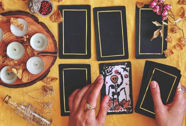 Wiccan witch reading the future at her altar using 5 card tarot spread on bright yellow cloth (flat lay) Hand painted Death tarot card with tarot deck in woman's hand with white candles and dried flowers and herbs in the background - Photo, Image