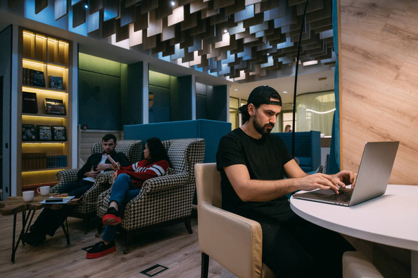 Männer und Frauen arbeiten im Coworking. Junger Mann mit Laptop bei der Arbeit online am Tisch und ein paar Kollegen plaudern, sitzt auf Sesseln in einem stilvollen Zimmer - Foto, Bild