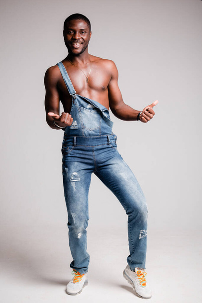Dark-skinned African-American man with naked torso in denim pants with suspenders crossed his arms on his chest. - Foto, imagen