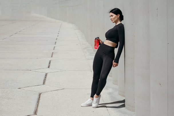 Beautiful fitness trainer in a black tight-fitting tracksuit stands near a concrete wall and holds a red drinking bottle outdoors in a park - Foto, Bild