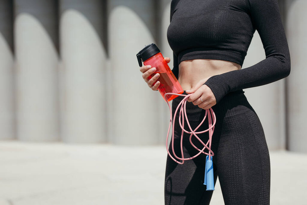 Close-up of the body of a girl athlete holding a jump rope and an orange drinking bottle for training outdoors in the park - Photo, image