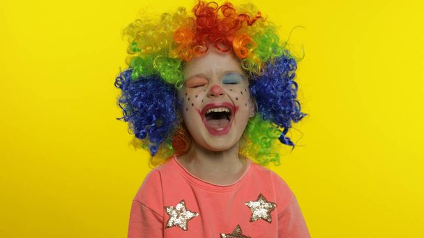 カラフルなウィッグの中で小さな子供の女の子ピエロは、楽しさ、笑顔、ダンスを持って、愚かな顔を作ります。ハロウィン - 写真・画像