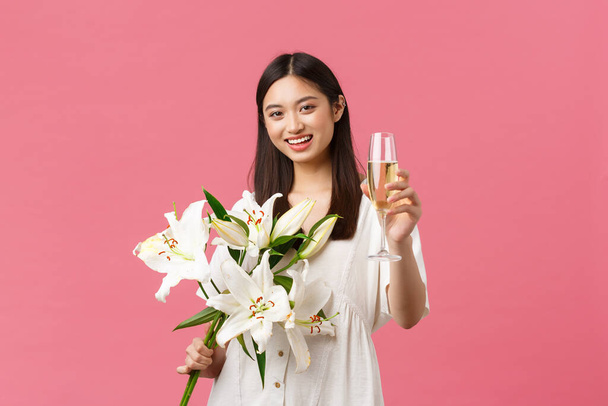 Feestdagen, feestdagen en leuk concept. Lachende mooie glamour aziatische vrouw in jurk met witte lelies boeket, het verhogen van glas champagne om toast te maken, drinken voor jarige meisje - Foto, afbeelding