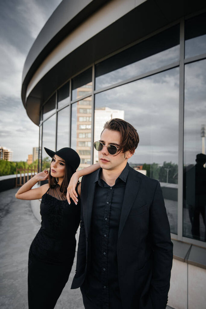 A beautiful, stylish pair of young people in black clothes and glasses stand against the background of an office building in the sunset. Fashion and style - Foto, immagini