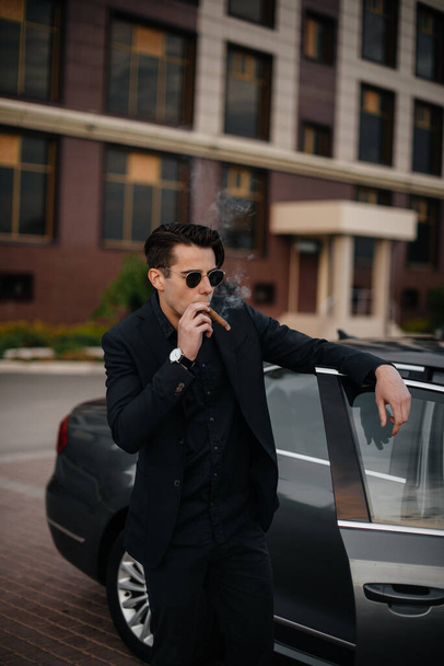 A stylish businessman smokes cigars near a luxury car. Fashion and business - Photo, Image