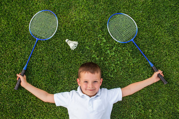 A smiling child holding two rackets in his hands lies on the grass next to the shuttlecock. Badminton game with a shuttlecock on the nature. Outdoor activity concept. - Photo, Image