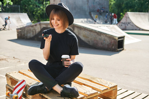Pretty smiling girl blogger communicate by phone sitting on wooden pallet in park with skateboard and cup coffee to go  - Foto, Imagen