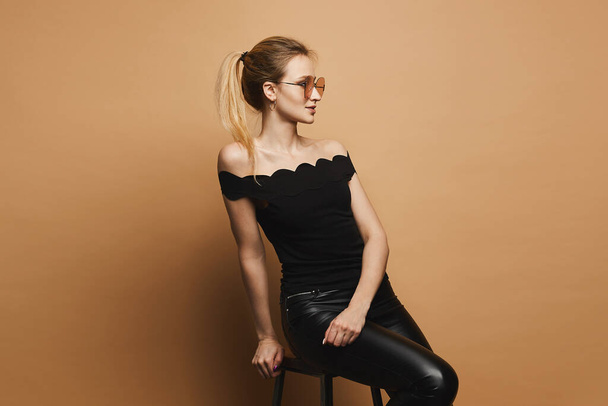 Beautiful and sexy blonde model girl with perfect sporty body and makeup in black leather pants, in black t-shirt and stylish sunglasses sits on stool and and posing at orange background - Foto, imagen