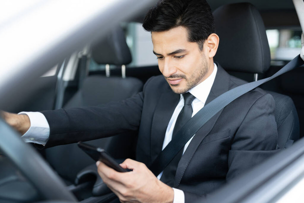 Hispanic young businessman using smartphone while driving car in city - Fotó, kép