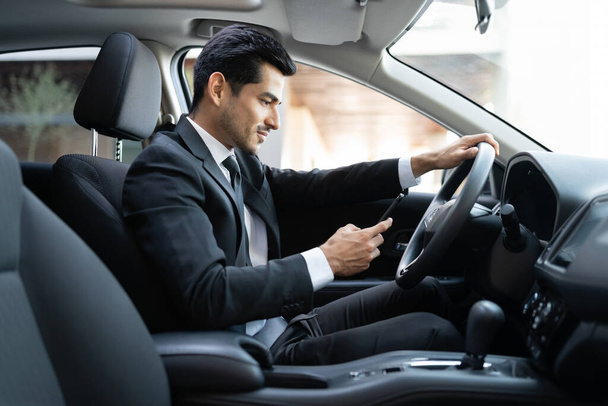 Уверенный бизнесмен, пользующийся смартфоном во время вождения автомобиля в городе
 - Фото, изображение