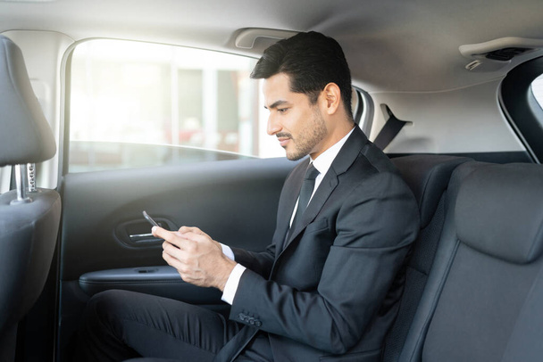 Widok z boku łacińskiego młodego biznesmena SMS za pośrednictwem smartfona podczas podróży taksówką - Zdjęcie, obraz