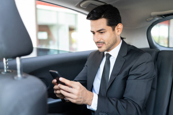 Hispanic young businessman text messaging through smartphone while traveling in taxi - Fotografie, Obrázek