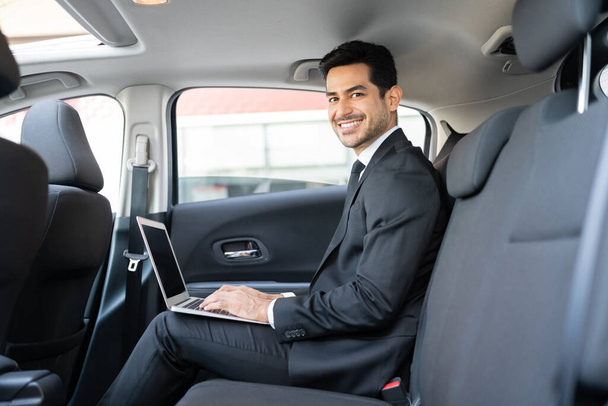 タクシーで旅行中にラップトップを使用しているヒスパニック系の若いビジネスマンの笑顔の肖像画 - 写真・画像