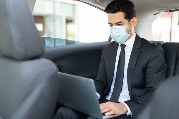 Businessman wearing face mask while using laptop in taxi during coronavirus outbreak - Fotó, kép