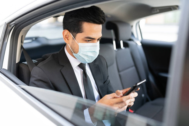 Empresário usando máscara facial enquanto usa smartphone em táxi durante crise coronavírus
 - Foto, Imagem