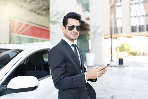 Zelfverzekerde jonge Latijnse zakenman met mobiele telefoon stand-by auto - Foto, afbeelding