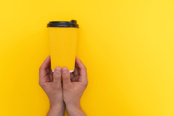 Two caucasian hands hang paper cup with lid on yellow background - 写真・画像