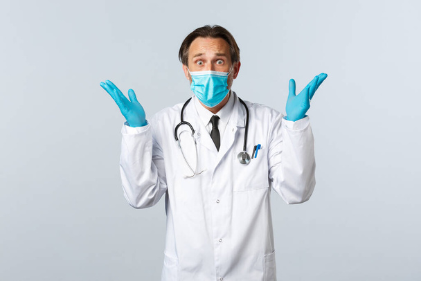 Covid-19, Prävention von Viren, Gesundheitspersonal und Impfkonzept. Alarmierte besorgte Ärztin in medizinischer Maske und Handschuhen, ängstlich starrend mit beunruhigten Händen nach oben, Panik über weißem Hintergrund - Foto, Bild