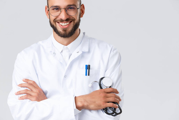 Healthcare workers, coronavirus, covid-19 pandemic and insurance concept. Close-up of handsome smiling male doctor in white coat, glasses, cross arms chest and hold stethoscope, talk to patients - Foto, Imagem