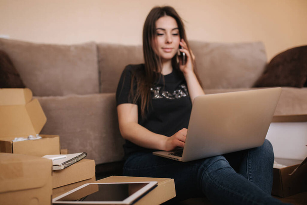 Šťastná mladá žena nakupuje online. Žena používá notebook a tablet pro hledání nového zboží. Atraktivní dívka obklopená balíčky - Fotografie, Obrázek