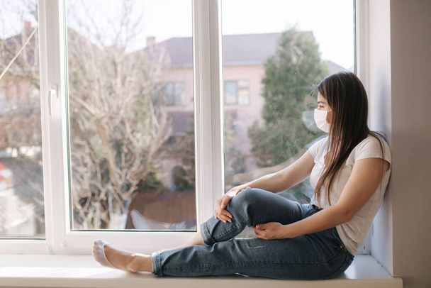 Πλευρική άποψη της νεαρής γυναίκας κάθεται στο περβάζι του παραθύρου και σε προστατευτική μάσκα και να κοιτάξουμε έξω από το παράθυρο. Μόνος στο σπίτι σε καραντίνα. Θέμα του Coronavirus. Μείνε σπίτι. - Φωτογραφία, εικόνα
