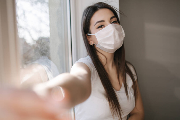 Attractive woman in medical mask make selfie at home. Female in white tshirt and white medical mask use phone - 写真・画像