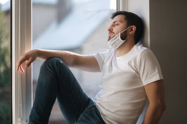 Young man sits on windowsill and hold in hands protective mask and looks out the window. Alone at home in quarantine. Coronavirus theme. Stay at home - Foto, Imagen