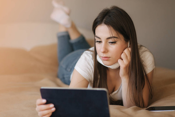 Młoda kobieta w masce używa tabletek na łóżku. Kobiety oglądają filmy online podczas kwarantanny. Studia studenckie w domu - Zdjęcie, obraz
