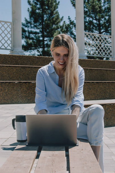 Η επιχειρηματίας μιλάει στο τηλέφωνο. Μια νεαρή επιχειρηματίας δουλεύει με ένα λάπτοπ με ένα μπλε πουκάμισο στο δρόμο σε ένα πάρκο κάτω από τον ήλιο! Επιχειρηματική έννοια - Φωτογραφία, εικόνα