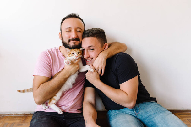 Foto d'archivio di due uomini omosessuali caucasici seduti a giocare con il loro gatto tabby arancione e bianco. - Foto, immagini