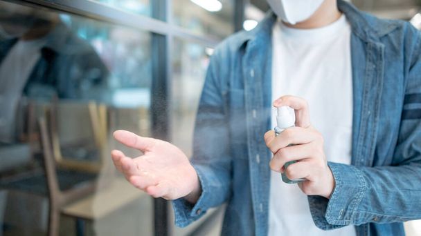 Καθαρισμός χεριών με απολυμαντικό σπρέι. Ασιάτης που φοράει μάσκα ψεκάζοντας αλκοόλ για την πρόληψη της λοίμωξης από τον ιό του κερατοειδούς (COVID-19) σε δημόσιο χώρο. Φροντίδα υγείας με αντιβακτηριδιακό προϊόν. - Φωτογραφία, εικόνα