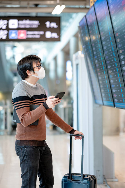 Aziatische man toeristische dragen gezichtsmasker controleren vlucht vanaf aankomst vertrekbord met behulp van smartphone in de terminal van de luchthaven. Coronavirus (COVID-19) pandemische preventie tijdens het reizen. Sociaal afstandconcept - Foto, afbeelding