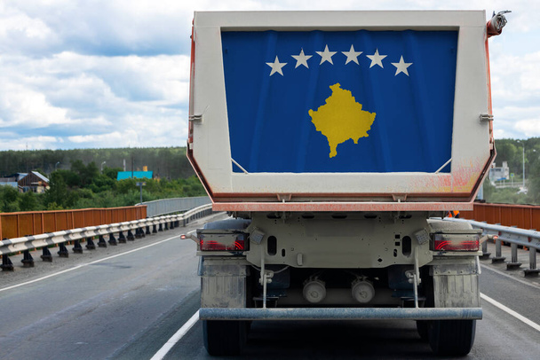 Gran camión sucio con la bandera nacional de Kosovo moviéndose en la carretera, en el fondo de la aldea y el paisaje forestal. Concepto de exportación-importación, transporte, entrega nacional de mercancías
  - Foto, Imagen