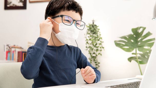 Online-Lernen & soziale Distanzierung, Netter asiatischer kleiner Junge setzt Kopfhörer auf, um aufgrund der Covid-19-Pandemie, Aussperrung und Schulunterbrechung zu Hause Online-Unterricht am Laptop zu lernen. - Foto, Bild