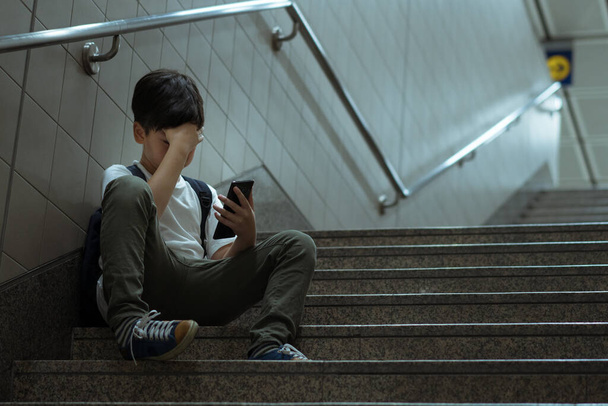 Cyber bullying concept. Young Asian preteen/teenage boy sitting at stair, covering his face with hand, other hand holding smartphone. Alone, stressed, frustrated, overwhelmed, crying and depressed - Photo, Image