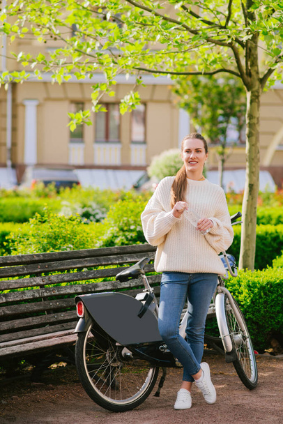 Girl in jeans and sweater standing near the bike in park - Fotoğraf, Görsel
