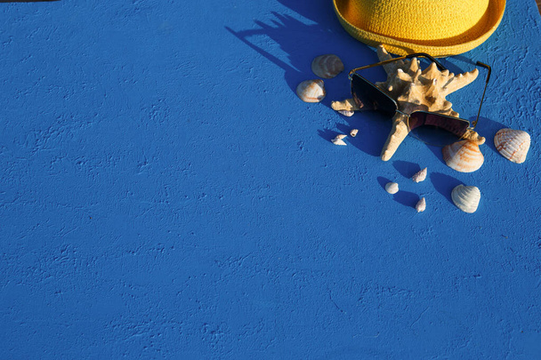 Frame with beach accessories on a nautical theme: yellow straw hat, sunglasses, starfish and shells on a blue background. Vacation concept, sea trip, UV protection, swimming. Copy space. Flatlay - Φωτογραφία, εικόνα